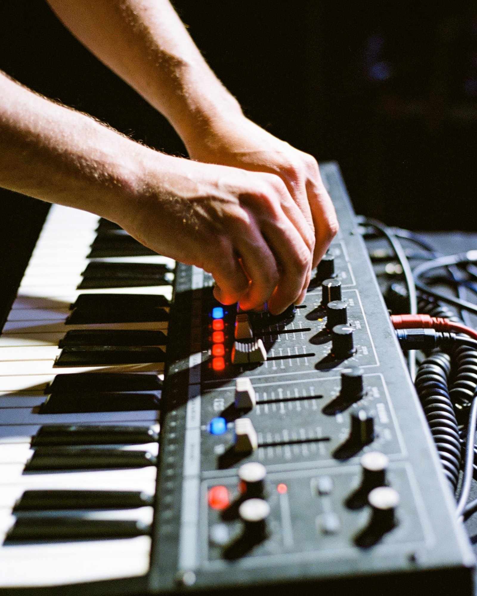 Hands adjusting knobs on a synthesizer lit by small red and blue lights on a dark stage