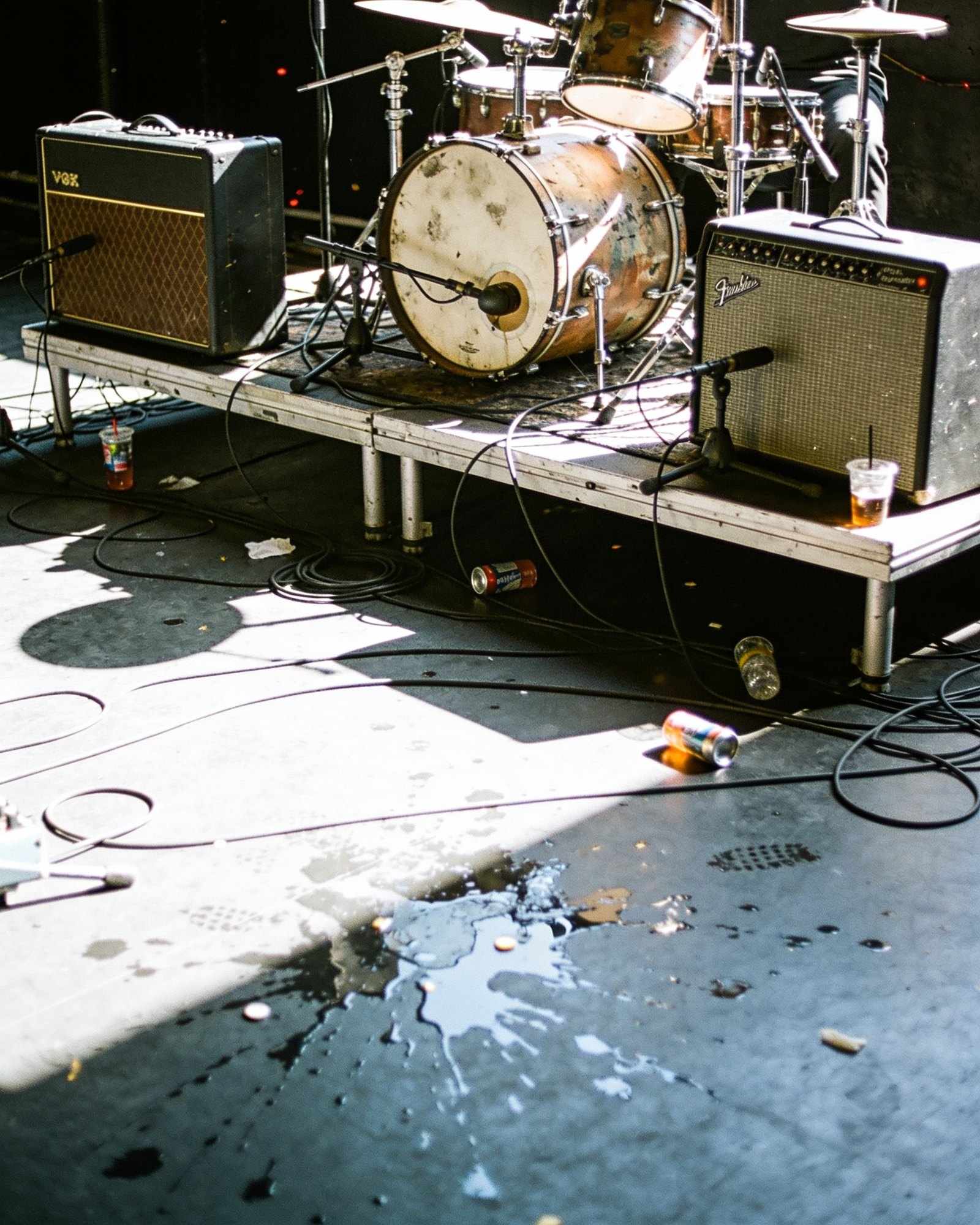 A battered drum kit and two guitar amplifiers on a small stage, cables everywhere, under hard white light