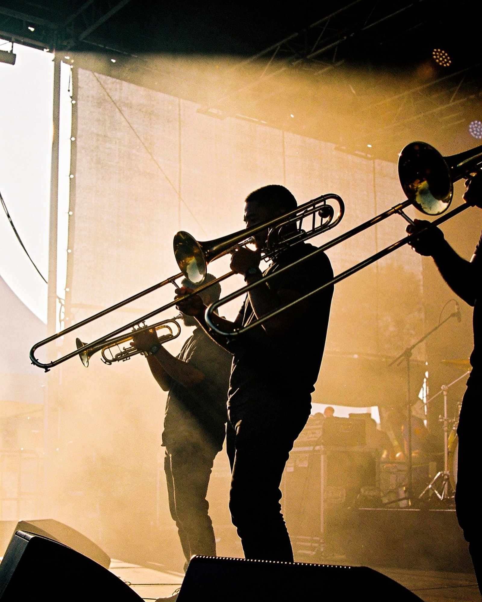 A brass section silhouetted against a wall of warm stage light and haze, seen from the side of the stage