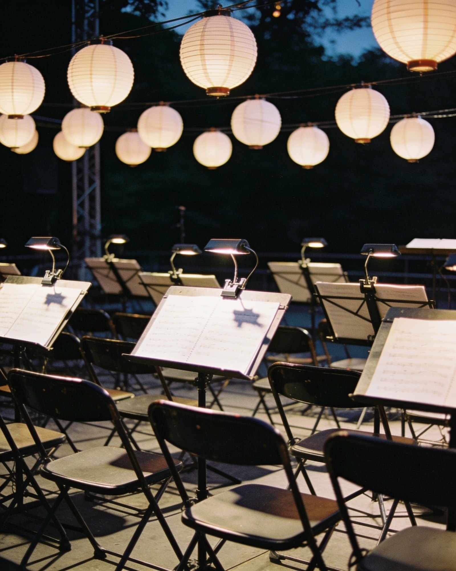 Rows of music stands with small lamps and sheet music on a darkening stage, paper lanterns strung overhead